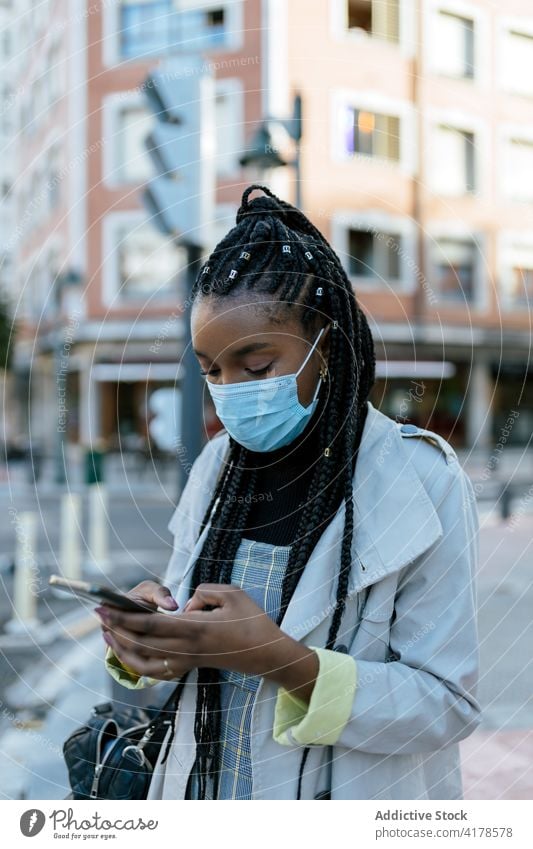 Schwarze Frau mit Maske benutzt Smartphone auf einer Straße in der Stadt Mundschutz Coronavirus urban benutzend Mobile Telefon Pandemie modern jung