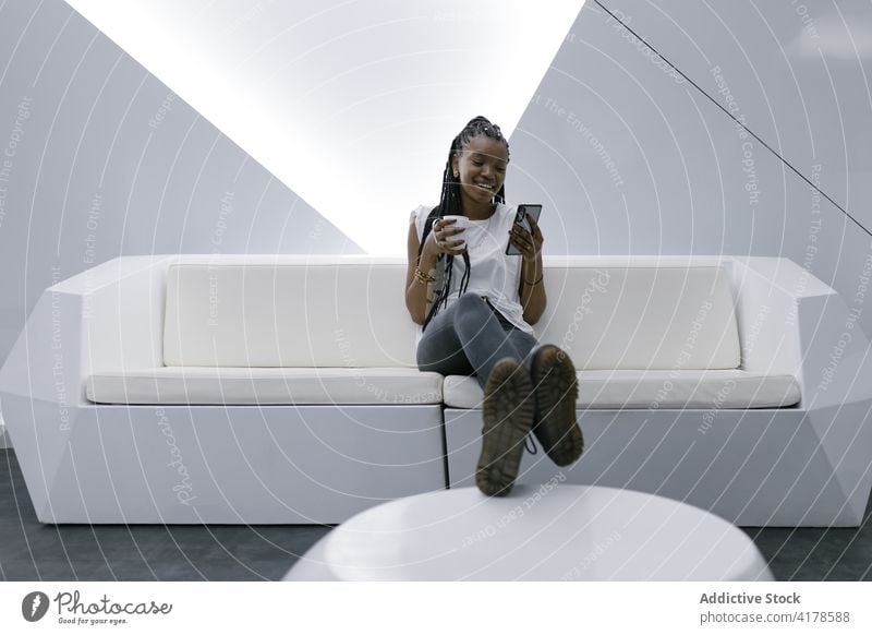 Fröhliche schwarze Frau, die sich in einem modernen weißen Wohnzimmer ausruht Browsen Smartphone futuristisch Innenbereich Geometrie Möbel heiter ethnisch