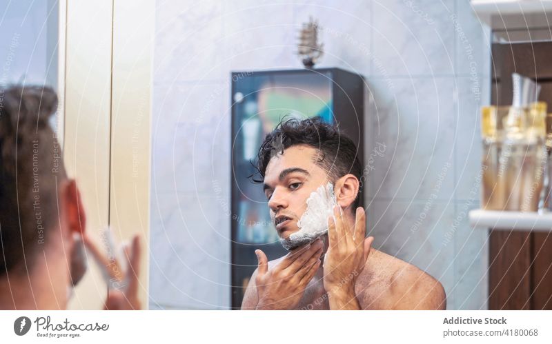 Mann rasiert Bart im Badezimmer Rasieren Vollbart Rasierer schäumen Routine Pflege Morgen jung männlich ethnisch brünett Spiegel ohne Hemd manuell Lifestyle