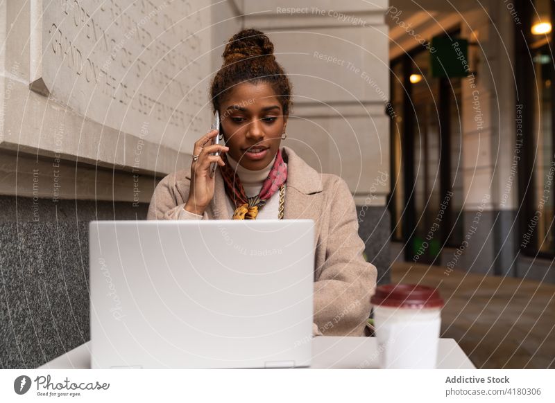 Intelligente junge schwarze Frau, die in einem Café mit ihrem Smartphone spricht und einen Laptop benutzt Telefonanruf Arbeit freiberuflich Stil klug Kaffee