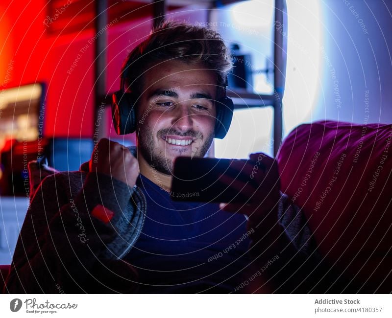 Positiver Mann mit Kopfhörern, der in einem dunklen Raum auf seinem Smartphone surft benutzend Dunkelheit Sofa Inhalt zuschauend Apparatur ruhen unterhalten