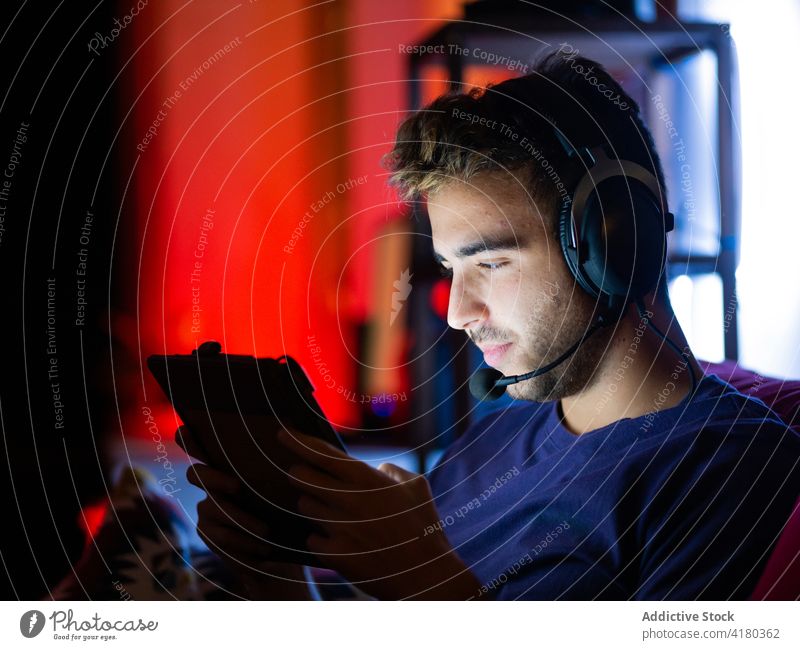 Positiver Mann mit Kopfhörern, der in einem dunklen Raum auf einem Tablet surft benutzend Dunkelheit Sofa Inhalt zuschauend Apparatur ruhen unterhalten spielen
