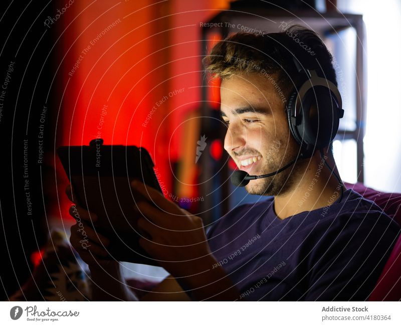 Positiver Mann mit Kopfhörern, der in einem dunklen Raum auf einem Tablet surft benutzend Dunkelheit Sofa Inhalt zuschauend Apparatur ruhen unterhalten spielen