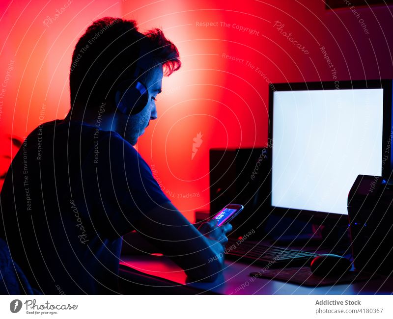 Ruhiger Mann, der ein Smartphone benutzt und am Tisch mit einem Computer sitzt benutzend Dunkelheit zuschauen Surfen Kopfhörer zuhören Gerät leerer Bildschirm