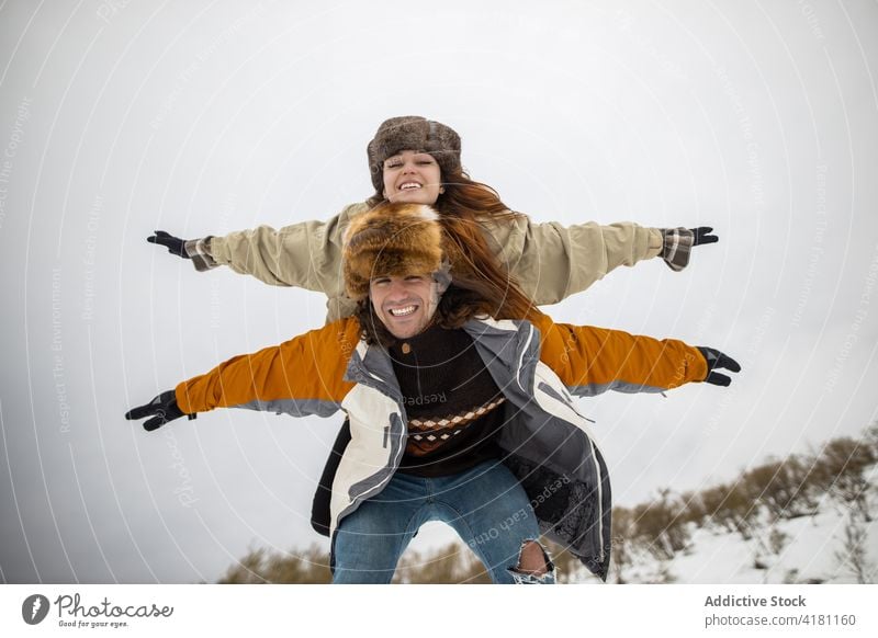 Glückliche Frau, die während eines Winterausflugs huckepack auf ihrem Freund reitet Huckepack Mitfahrgelegenheit Freundin Arme hochgezogen Freiheit Spaß haben