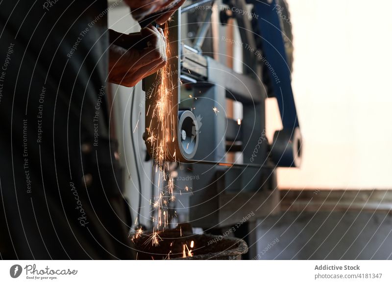 Unbekannter männlicher Schmied, der ein Eisenteil von Hand mit einer Maschine in einer Schmiede schleift Mann polnisch Schleifmaschine Metallbearbeitung