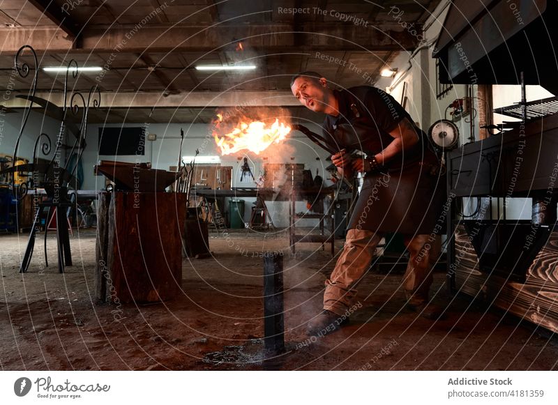 Aufmerksamer junger Schmied, der beim Erhitzen einer Zange in der Werkstatt das Feuer anbläst Mann erwärmen Schmiede Schlag Flamme Konzentration Arbeit