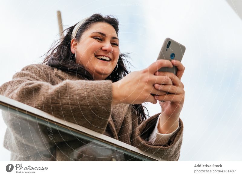 Lächelnde Frau, die in der Nähe eines städtischen Zauns eine Textnachricht auf ihrem Smartphone schreibt plaudernd Internet online freie Zeit Großstadt