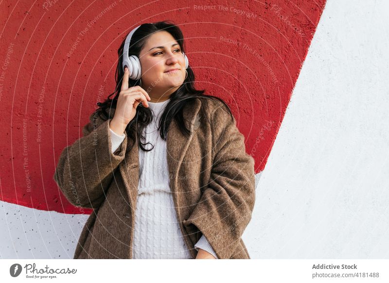Lächelnde Frau hört Musik über Kopfhörer an einer hellen Wand zuhören Drahtlos verträumt genießen meloman Straße benutzend Apparatur Gerät bewundern Inhalt