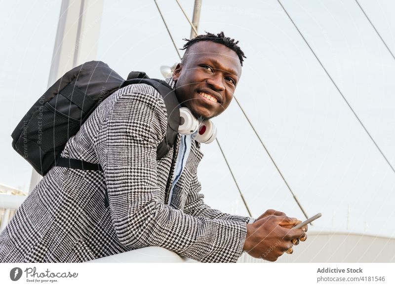 Glücklicher schwarzer Büroangestellter mit Smartphone auf Hängebrücke Manager Lächeln Kopfhörer formal Rucksack nach vorne lehnen Brücke Mann Stadt Apparatur