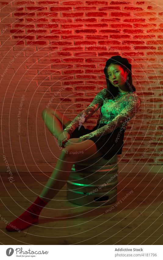 Trendy asiatische Frau sitzt auf Metallfass in dunklem Studio Stil selbstbewusst Anmut trendy Lauf Outfit Atelier cool Vorschein Streetstyle sitzen