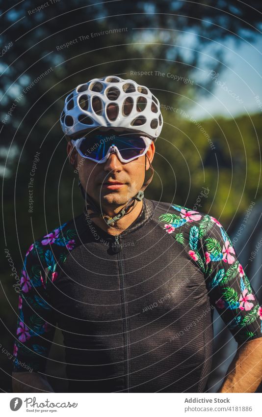 Seriöser Sportler in Radfahrerkleidung steht in der Nähe von grünen Bäumen im Sonnenlicht Mann selbstbewusst Athlet Park Training Porträt Baum ernst Aktivität