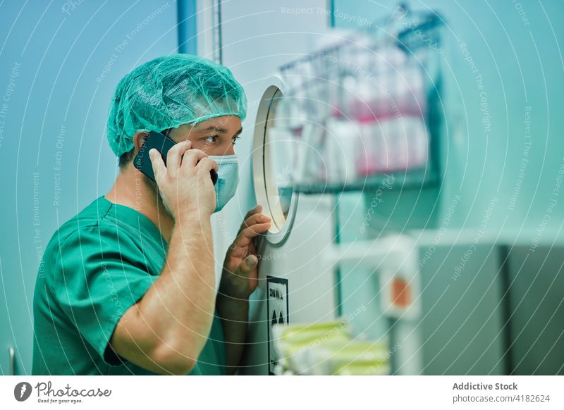 Fokussierter männlicher Sanitäter, der während einer Operation mit seinem Smartphone spricht und durch ein Fenster schaut Mann reden Arzt Operationssaal