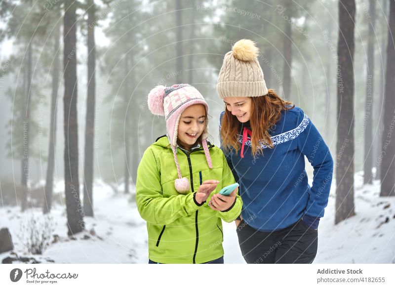 Fröhliches Mädchen und Mutter mit Smartphone im Winterwald Frau benutzend Wald Spaziergang heiter Zahnfarbenes Lächeln Oberbekleidung Tochter Glück Waldgebiet