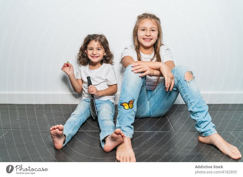 Fröhliche kleine Schwestern sitzen auf dem Boden Mädchen heiter Glück Stock niedlich Kindheit Zusammensein Freude zu Hause Zahnfarbenes Lächeln genießen