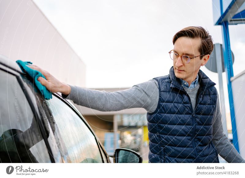 Konzentrierter junger Mann, der sein Auto mit einer Serviette abwischt PKW Waschen Wischen Tuch Fokus Fahrzeug Selbstbedienung Station Straße Fenster Wasser