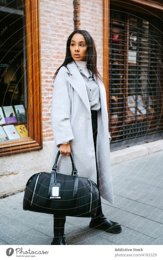 Stilvolle Frau im Mantel auf der Straße stehend trendy Graben modern urban Accessoire Tasche Straßenbelag Bürgersteig Lifestyle Zeitgenosse ethnisch Outfit