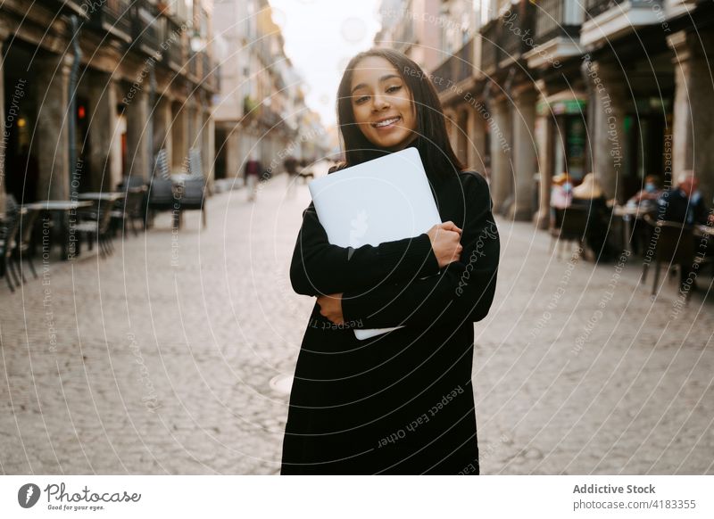 Fröhliche Frau mit Laptop auf sonniger Straße heiter urban Schüler Fußgänger Stadt aufgeregt Netbook tragbar Sonnenlicht Stil Gerät Lächeln Zeitgenosse ethnisch