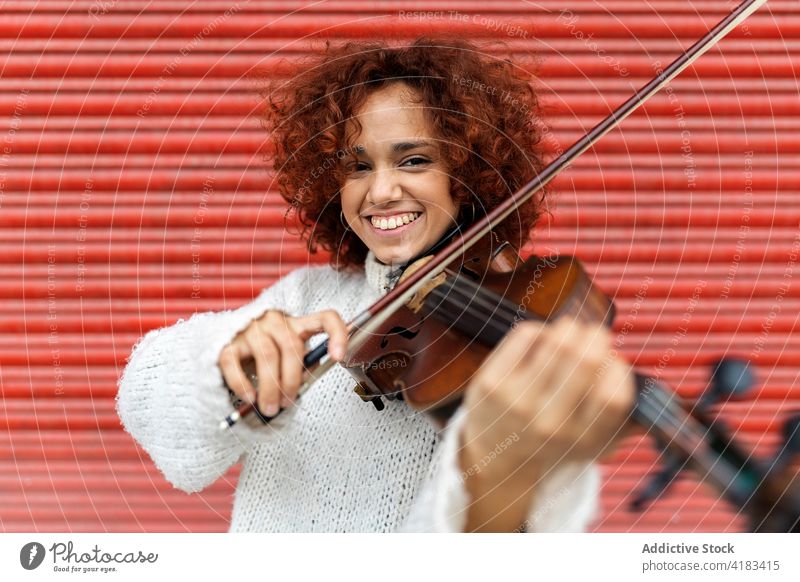 Fröhliche rothaarige Frau spielt Geige vor roter Wand spielen Musiker Glück Zahnfarbenes Lächeln Instrument akustisch ausführen live Melodie Talent Fähigkeit
