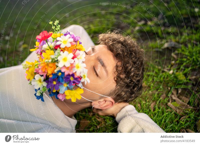 Unbekannter Mann mit Blumenmaske im Gesicht auf einer Wiese ruhend Mundschutz COVID Konzept Augen geschlossen Blüte hell sich auf die Hand lehnen Bund 19