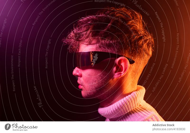 Trendiger junger Mann mit futuristischer Sonnenbrille in einem Neonstudio ausrichten Stil Mode selbstsicher neonfarbig charismatisch Persönlichkeit modern