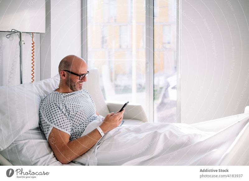 Glücklicher männlicher Patient, der auf dem Stationsbett telefoniert Mann geduldig Bett Smartphone benutzend Telefon Mobile Fokus Arbeitstier Gerät Krankenhaus
