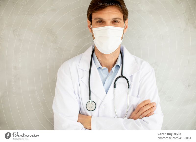 Attraktiver junger männlicher Arzt mit chirurgischer Maske und an der Wand stehend medizinisch Pandemie Virus Mundschutz Krankheit Coronavirus Gesundheit