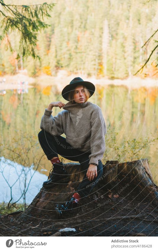 Stilvoller Reisender auf Baumstumpf gegen See im Herbst Tourist Gesicht berühren cool ruhen Frau Stumpf Reflexion & Spiegelung Natur anhaben stylisch Ausflug
