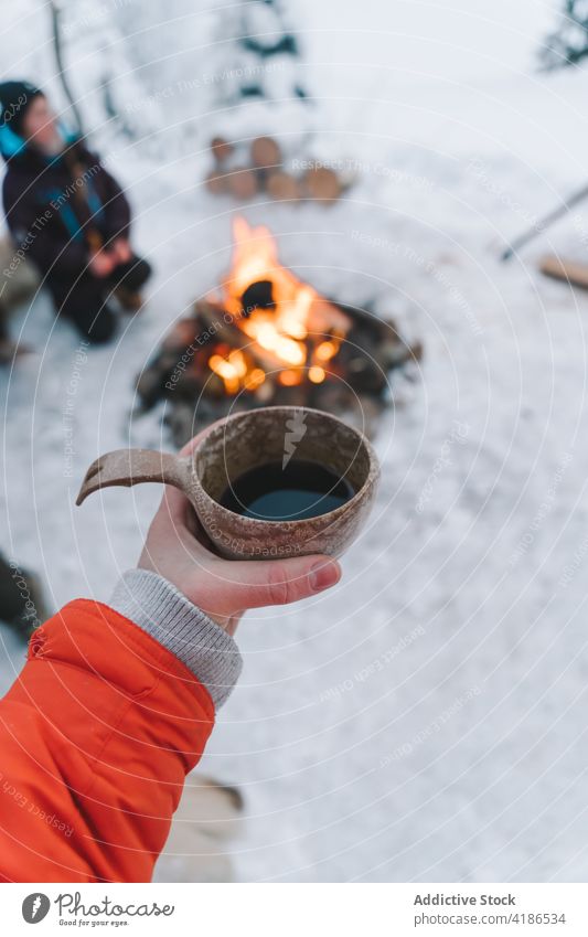 Unbekannte Frau trinkt heißes Getränk am Lagerfeuer an einem verschneiten Wintertag trinken Kaffee Schnee Heißgetränk Freudenfeuer Campingplatz Ausflug Urlaub