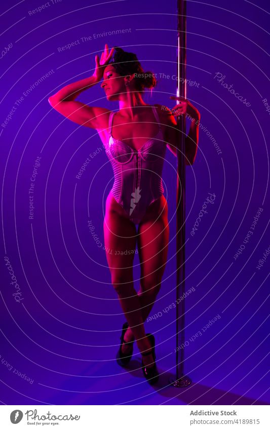 Anmutige Tänzerin berührt Kopf in der Nähe Tanzstange in Neonlicht Tanzen Mast Tastkopf Beine gekreuzt Körper Augen geschlossen erotisch neonfarbig Frau