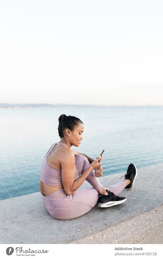 Ethnische Sportlerin surft im Internet auf dem Smartphone gegen das Meer auf dem Damm MEER online Pause Natur Beine gekreuzt Stauanlage benutzend Apparatur Frau