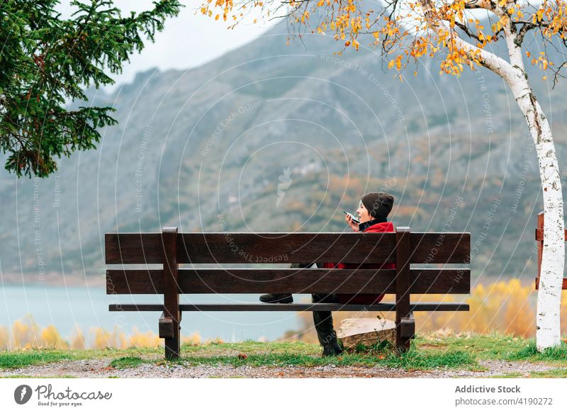 Frau sitzt auf einer Bank in einem majestätischen Herbstpark bewundern sprechend Smartphone Telefon Mobile Park Berge u. Gebirge Seeufer ruhen genießen