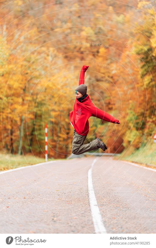 Glückliche Frau in Oberbekleidung, die im Herbst auf einer Landstraße springt springen aufgeregt Arme hochgezogen Wald in der Luft Straße Freiheit Freude