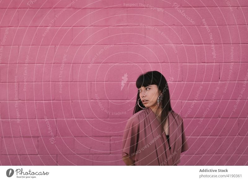 Modische junge ethnische Dame steht in der Nähe von rosa Wand und schaut weg Frau besinnlich nachdenklich Stil cool Mode Windstille Straße verträumt traumhaft