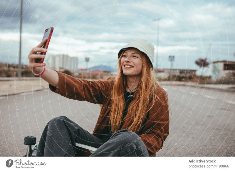 Zufriedene junge Frau sitzt auf dem Skateboard und nimmt Selfie in der Landschaft Lächeln Smartphone Freude Stil soziale Netzwerke tausendjährig Mobile Telefon