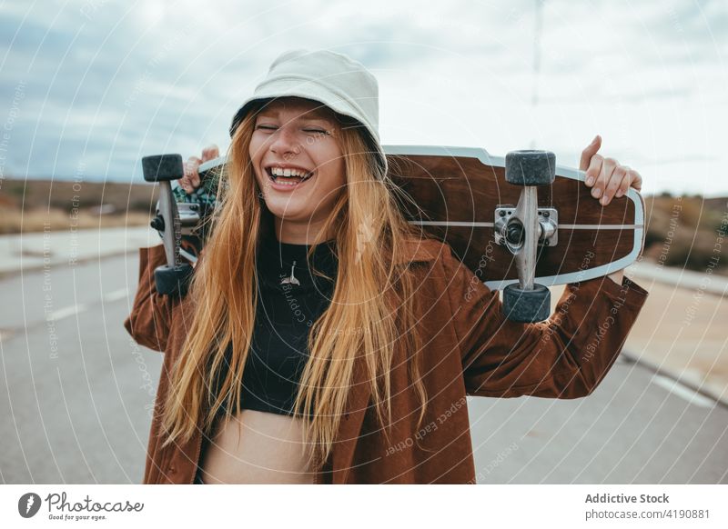 Fröhliche junge Frau lacht, während sie mit einem Skateboard in den Händen auf der Straße steht Lachen Augen geschlossen Longboard Aktivität selbstsicher heiter