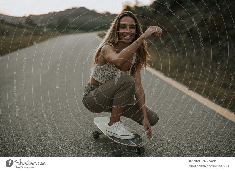 Fröhliche Skaterin, die sich auf einer Landstraße auf ein Cruiser Board kauert Frau sorgenfrei heiter Aktivität Mitfahrgelegenheit Voute Straße ländlich