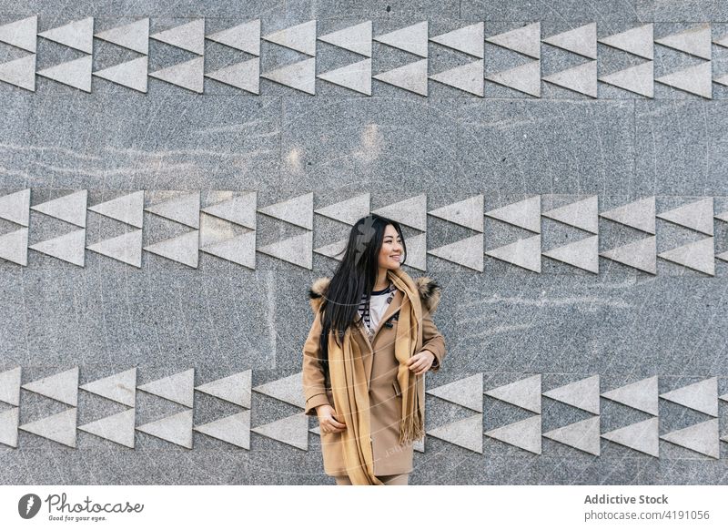 Lächelnde asiatische Frau in Mantel und Schal steht in der Nähe eines Gebäudes positiv Individualität Ornament Wand Vorschein Geometrie sorgenfrei heiter