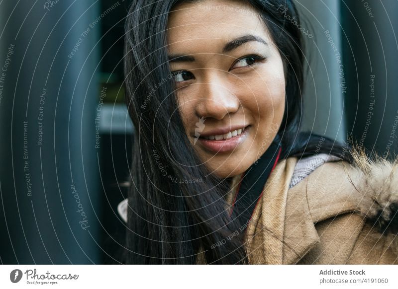 Lächelnde asiatische Frau in Mantel und Schal steht in der Nähe eines Gebäudes positiv Individualität Vorschein Geometrie sorgenfrei heiter Optimist