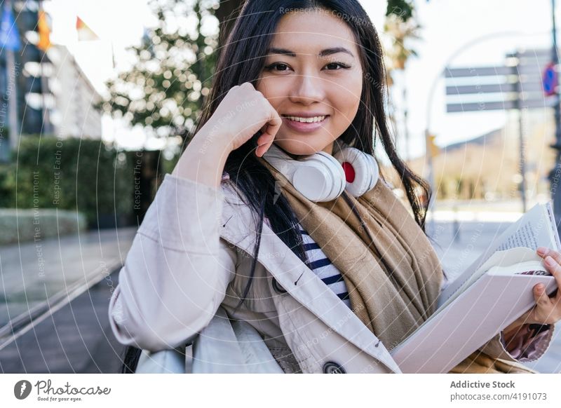 Lächelnde asiatische Frau, die auf einer Bank im Park sitzt und ein Buch liest lesen Optimist Etage Literatur Hobby Kopfhörer Drahtlos Belletristik Wissen