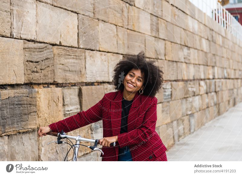 Erfreute ethnische Frau mit Fahrrad in der Stadt Spaziergang Großstadt Straße heiter Lächeln urban Fahrzeug schwarz Afroamerikaner alt Stein Wand Gebäude