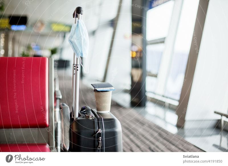 Neben den Sitzen in einem leeren Flughafen steht ein Koffer, darauf steht ein Glas Kaffee, daran hängt eine medizinische Schutzmaske. Coronavirus, soziale Distanz, Isolation