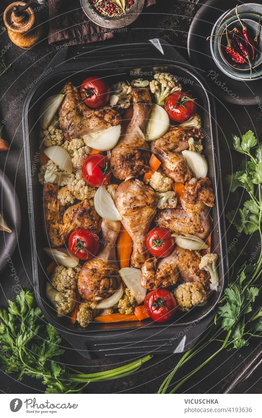 Leckere Hähnchenkeulen mit Gemüse in einer gusseisernen Pfanne auf dunklem, rustikalem Hintergrund mit frischen Kräutern. Gesundes Diät-Essen. Ansicht von oben. Hühnerkasserolle