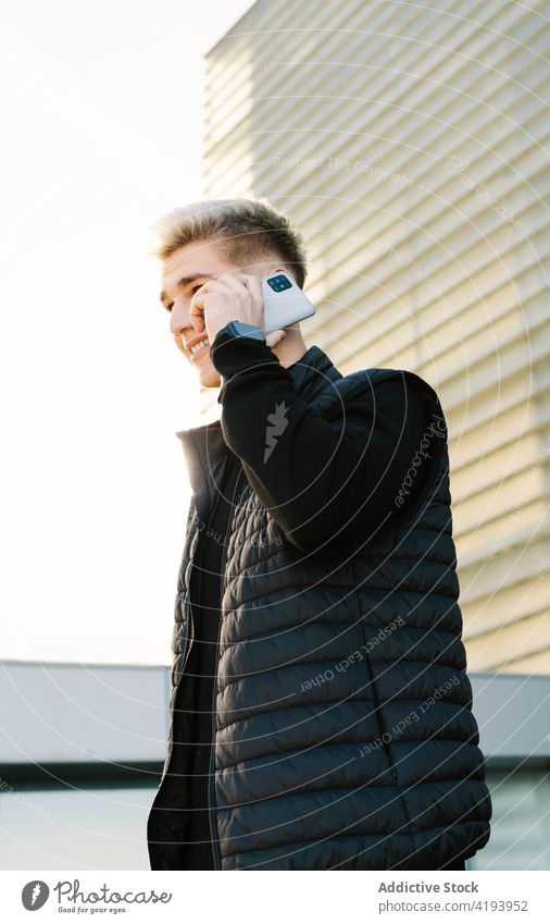 Positiver junger Mann, der mit seinem Smartphone spricht und in der Nähe eines modernen Gebäudes lächelt reden Straße Lächeln Telefonanruf Gespräch