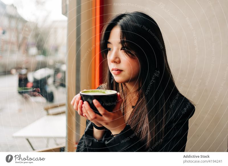 Langhaarige brünette asiatische Frau, die in einem Café einen Kaffee trinkt, während sie durch das Fenster auf ein Handy schaut Menschen Werkstatt Chinesisch
