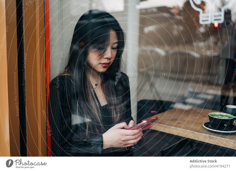 Langhaarige, brünette, asiatische Frau, die in einem Café einen Kaffee trinkt, während sie ein Handy betrachtet Funktelefon Telefon Kaffeehaus Chinesisch Mobile