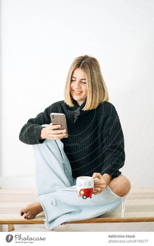 Lächelnde Frau mit heißem Getränk, die zu Hause mit ihrem Smartphone plaudert plaudernd Heißgetränk Internet online gemütlich heimwärts benutzend Apparatur