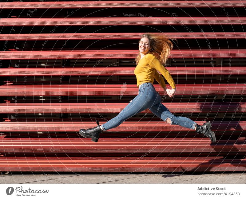 Fröhliche junge Frau springt auf der Straße im Sonnenlicht springen Lächeln Energie Freude heiter aktiv Inhalt Optimist Glück lässig tausendjährig Metall Zaun