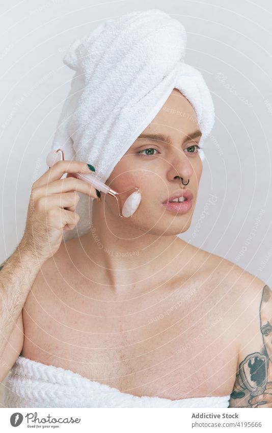 Queer massiert Gesicht mit Walze auf weißem Hintergrund queer Massage Rolle Gesichtsbehandlung Schönheit natürlich Tattoo Angebot beschaulich Porträt Mann