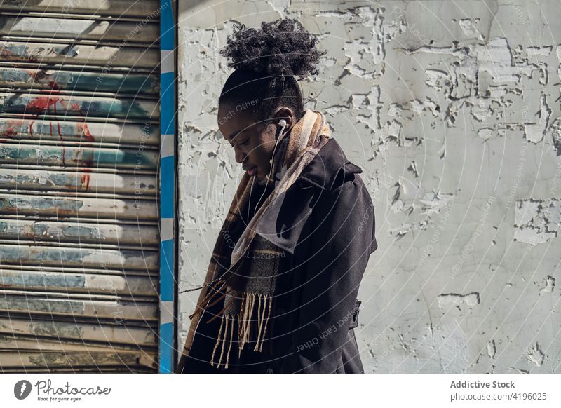 Schwarze Frau hört auf der Straße Musik aus Kopfhörern zuhören Gesang Audio feminin achtsam benutzend Gerät Stadt stylisch charmant urban Wand Kleidungsstück